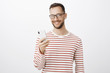 © Cookie Studio - Indoor shot of satisfied carefree entrepreneur in black glasses, talking on smartphone via wireless earphones and enjoying great quality of new gadget sound, standing over gray background