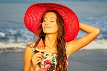 Beautiful And Young Girl In A Red Straw Hat Holds A Piece Of Watermelon And Enjoys A Vacation On The Beach. Red Juicy Watermelon On A Hot Summer Day Refreshing