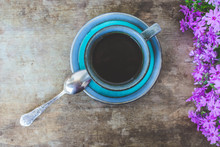 Top View Of An Blue Vintage Cup Of Black Coffee And Pink Or Purple Small Flowers On An Old, Rustic Wooden Table. Good Morning Coffee, Breakfast Or Floral Spring Background