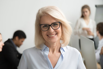 Wall Mural - Portrait of smiling senior businesswoman wearing glasses with businesspeople at background, happy older team leader, female aged teacher professor or executive woman boss looking at camera, head shot