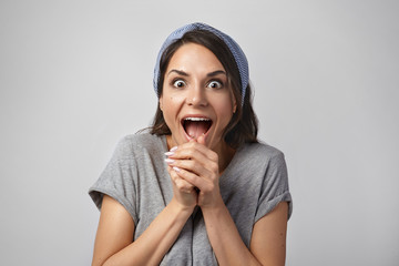 Tell me more. Studio shot of curious bug eyed young dark haired woman clasping hands and shouting with excitement and astonishment, opening mouth widely, listening to unexpected positive news