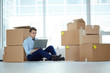 © pressmaster - Young serious businessman with laptop sitting by stack of boxes on the floor by large window and networking