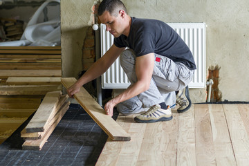 Young handsome professional carpenter installing natural wooden planks on wooden frame floor in empty unfinished room under reconstruction. Improvement, renovation and carpentry concept