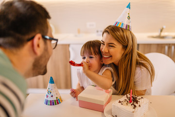 Wall Mural - Cute little girl is with her parents in their home and she is blowing a party horn because it is her birthday