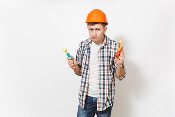 Young strange handsome man in casual clothes and protective orange hardhat holding toy hammer and saw isolated on white background. Instruments for renovation apartment room. Repair home concept.