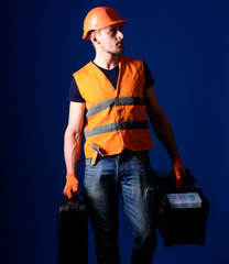 Man in helmet, hard hat holds toolbox and suitcase with tools, blue background. Professional repairman concept. Worker, handyman, repairman, builder on calm face carries bags with professional tools.