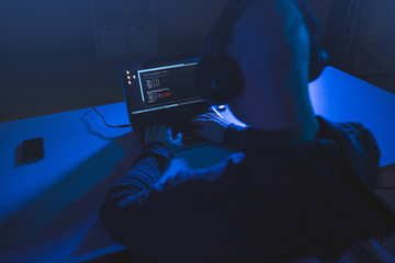 cybercrime, hacking and technology concept - male hacker with headphones and coding on laptop computer screen wiretapping or using computer virus program for cyber attack in dark room