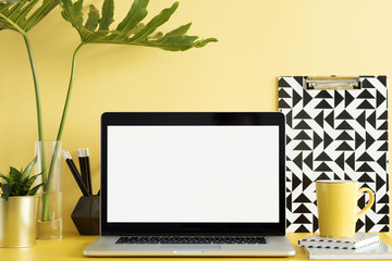 Stylish office workspace with laptop mock up screen, office accessories, leafs and plants on the yellow wall. Space of freelancer web developer.