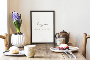 Stylish interior design of kitchen space with small table with mock up frame, plant, cups of tea and tasty dessert.