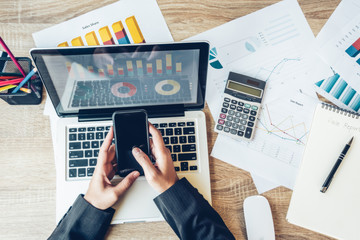 Business women using phone on workplace with laptop. close up hands on a keyboard for and financial data analyzing counting. Top view,Workplace strategy Concept.