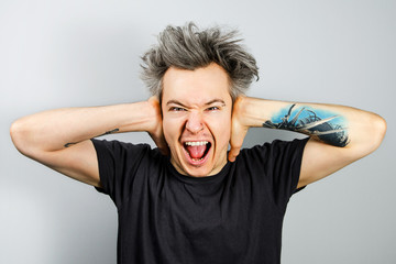 Unshorn and unshaven young guy with piercings on his face, closing his ears with his hands and opening mouth, on gray background.