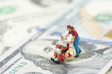 Cost of retirement living, health insurance or medical industry business concept, miniature senior old woman figurine sitting on wheelchair on US dollar banknotes money