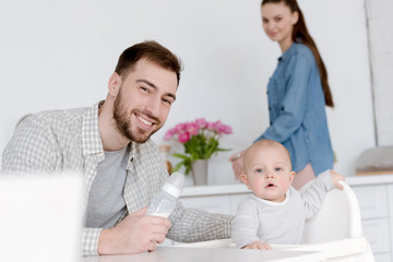Wall Mural - father feeding son with milk in baby bottle, mother standing on kitchen behind
