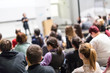 © kasto - Female speaker giving presentation in lecture hall at university workshop. Audience in conference hall. Rear view of unrecognized participant in audience. Scientific conference event.