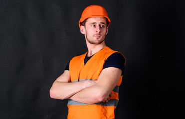 Man in helmet, hard hat hold arms crossed on chest, black background. Worker, contractor, builder on strict face with muscular biceps. Builder in helmet posing, copy space. Strong builder concept.