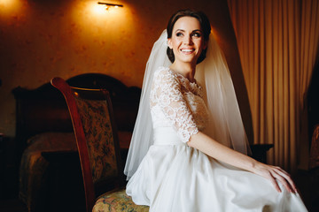 Beautiful brunette bride woman in wedding dress and long white veil sitting on the luxury vintage chair in dark room and smiling sincerely.