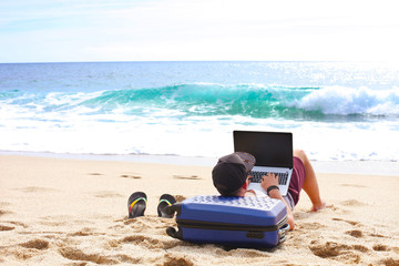 Young man, programmer on sandy beach leaning on suitcase, coding on a blank screen laptop computer, typing, blogging. Freelance, distance work concept. Sea view, ocean waves. Background, copy space
