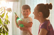 © sementsova321 - Plant care concept. Cheerful little male kid holds spray bottle, wants to water flower, stands neear his affectionate mother near window at home. Young mum and her son spray plant indoor together
