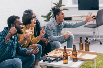 Wall Mural - multicultural young friends sitting on couch near table with pizza and beer bottles while watching american football match