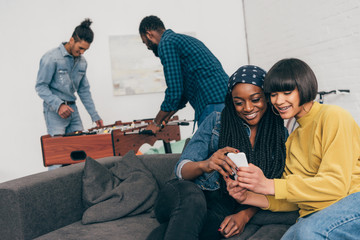 Wall Mural - two young female friends sitting with smartphone and two young men playing table soccer behind