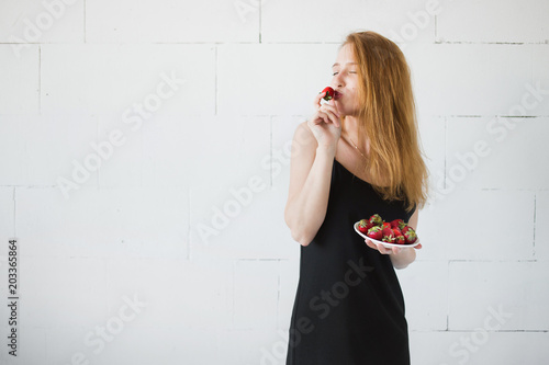 Fotografie, Obraz Young sexy slim woman eating sweet juicy strawberry near white wall