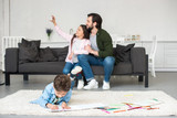 Fototapeta Nowy Jork - happy father and daughter sitting on sofa and taking selfie with smartphone while little boy drawing on carpet at home