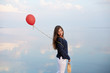 Minimalistic portrait of young woman with red air balloon and present bag near the calm sea or lake shore. Clouds reflected on smooth water surface. Girl on her birthday. Copyspace. Holiday concept.