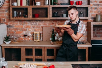 Wall Mural - Young man proud of himself standing at kitchen with freshly baked pizza. Homemade food, cooking man, people concept