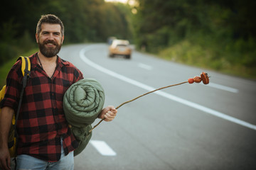 Wall Mural - Bearded smiling man with rucksack, sleeping back holding stick with sausages on it. Handsome hitchhiker on the way back home, wanderlust and adventure concept