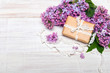 Prezent z kokardą otoczony kwiatami bzu na białym drewnianym tle, miejsce na tekst. Świąteczna kompozycja z okazji Dnia Matki, urodzin lub rocznicy. 