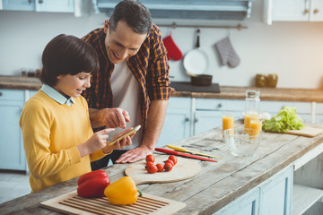 Father and son standing at the kitchen table. They are looking at mobile phone with joy. Vegetables are on desk. Copy space in right side