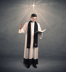 A young male priest in black and white giving his blessing in front of grey wall with glowing cross concept.