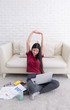© weedezign - Asian female arm up to stretch oneself after use laptop computer to work in living room at home.woman sit back at sofa on carpet at brick wall working online with data sheet and book..
