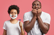 © WHstudio Leushin N - Photo of pleased handsome teenage boy with foam on face, holds razor, teaches to shave and nervous young African American male bite finger nails, look anxious, isolated over pink studio background
