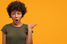 Photo Of Greatly Surprised Dark Skinned Female With Bushy Afro Haircut, Wears Casual T Shirt, Indicates At Yellow Blank Copy Space, Keeps Mouth Widely Opened, Being Astonished With Something.