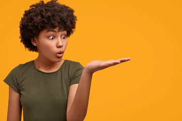 Wall Mural - Horizontal shot of surprised African American woman indicates with palm away, shows something unexpected, stares with shock, isolated over bright yellow background. People, emotions, advertisement
