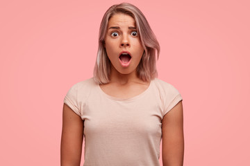 Wall Mural - Image of frightened young European pretty female has wide opened mouth, stares at camera with bugged eyes, being scared by someone, expresses her big surprisement and astonishment, dressed casually.