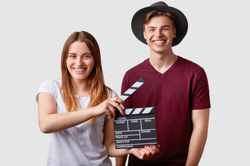 Two successful young female and male famous producers or directors hold film clapper, participate in shooting film, have joyful expressions, pose against white background. Film making concept.