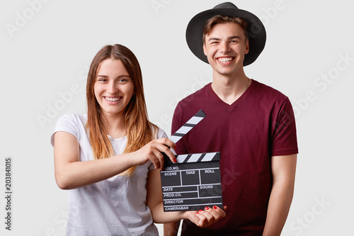 Fotografie, Obraz Two successful young female and male famous producers or directors hold film clapper, participate in shooting film, have joyful expressions, pose against white background