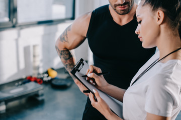 female personal trainer writing on clipboard and sportsman standing near at gym