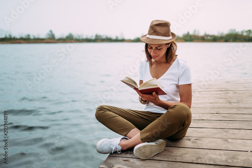 Cuadros en Lienzo Portrait of a girl reading a book while sitting on a small wooden wharf