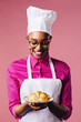 © Carlos David - Portrait of a happy young woman in cooking hat and apron looking at freshly made croissant, isolated on pink studio background