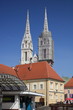 Centrum Zagrzebia, stolicy Chorwacji, z budynkiem zadaszonym pasażem oraz widocznymi zza budynku dwiema wieżami katedry katolickiej, jedna z wież restaurowana, z rusztowaniem, błękitne niebo