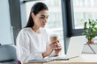 © LIGHTFIELD STUDIOS - young businesswoman holding paper cup and using laptop at workplace