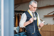 © LIGHTFIELD STUDIOS - handsome tailor standing with cup of coffee and looking at smartphone at sewing workshop