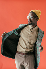 Wall Mural - fashionable african american man posing in vintage glasses and jacket, isolated on red