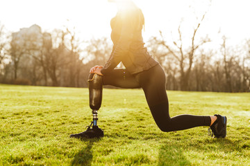 Wall Mural - Cropped image of disabled athletic girl in black sportswear, doing lunges and stretching prosthetic leg on grass