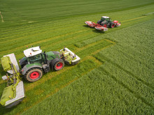 Aerial View Of Two Tractor Mowing A Green Fresh Grass Field, 
 Farmer In A Modern Tractors Mowing A Green Fresh Grass Field On A Sunny Day