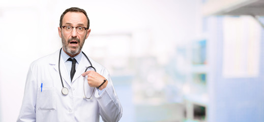 Poster - Doctor senior man, medical professional happy and surprised cheering expressing wow gesture, pointing with finger at hospital