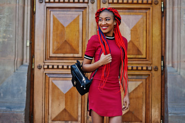 Wall Mural - Cute and slim african american girl in red dress with dreadlocks and backpack posed against school large wooden door. Stylish black student.